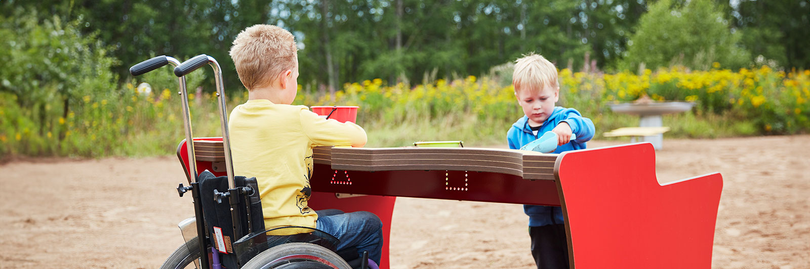 Children play at an outdoor playground sandtable which is elevated. One of the children is a wheelchair user.